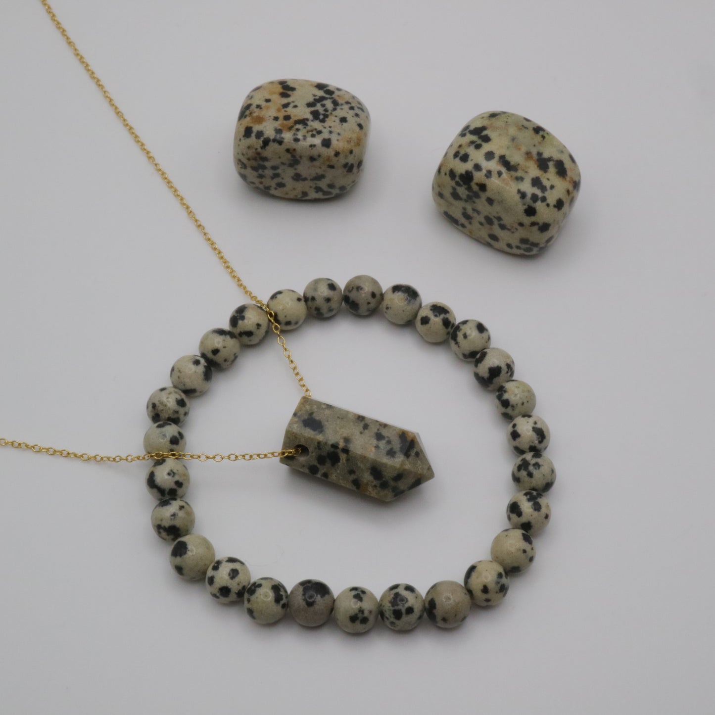 Dalmatian Jasper Crystal on a fine gold chain surrounded by a Dalmatian Jasper crystal bracelet and two Dalmatian Jasper tumblestone crystals